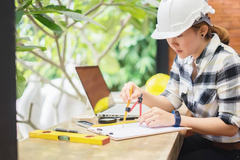 Female wearing a hard hat working at a desk with computer and other tools.